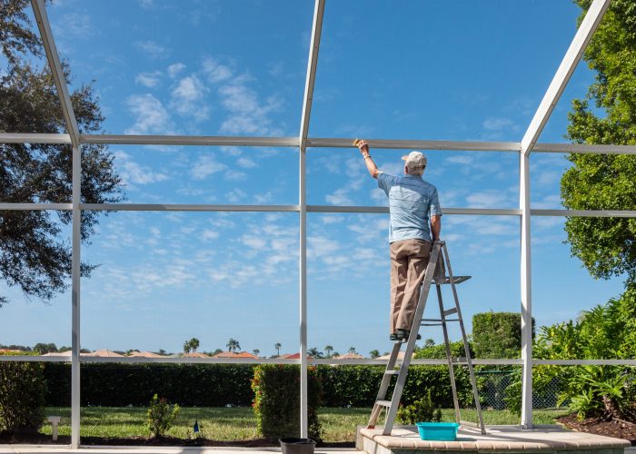 Handyman on ladder cleaning outdoor pool cage enclosure. Screened swimming pool lanai maintenance and screen repair.